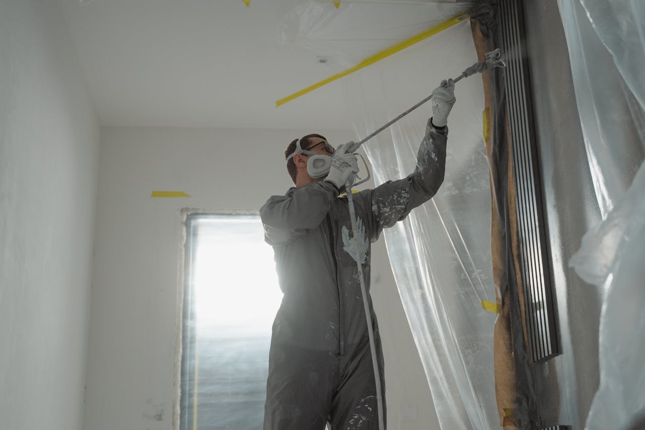 why-choose-us A professional painter wearing protective gear spray paints a wall indoors, working on a home renovation project.
